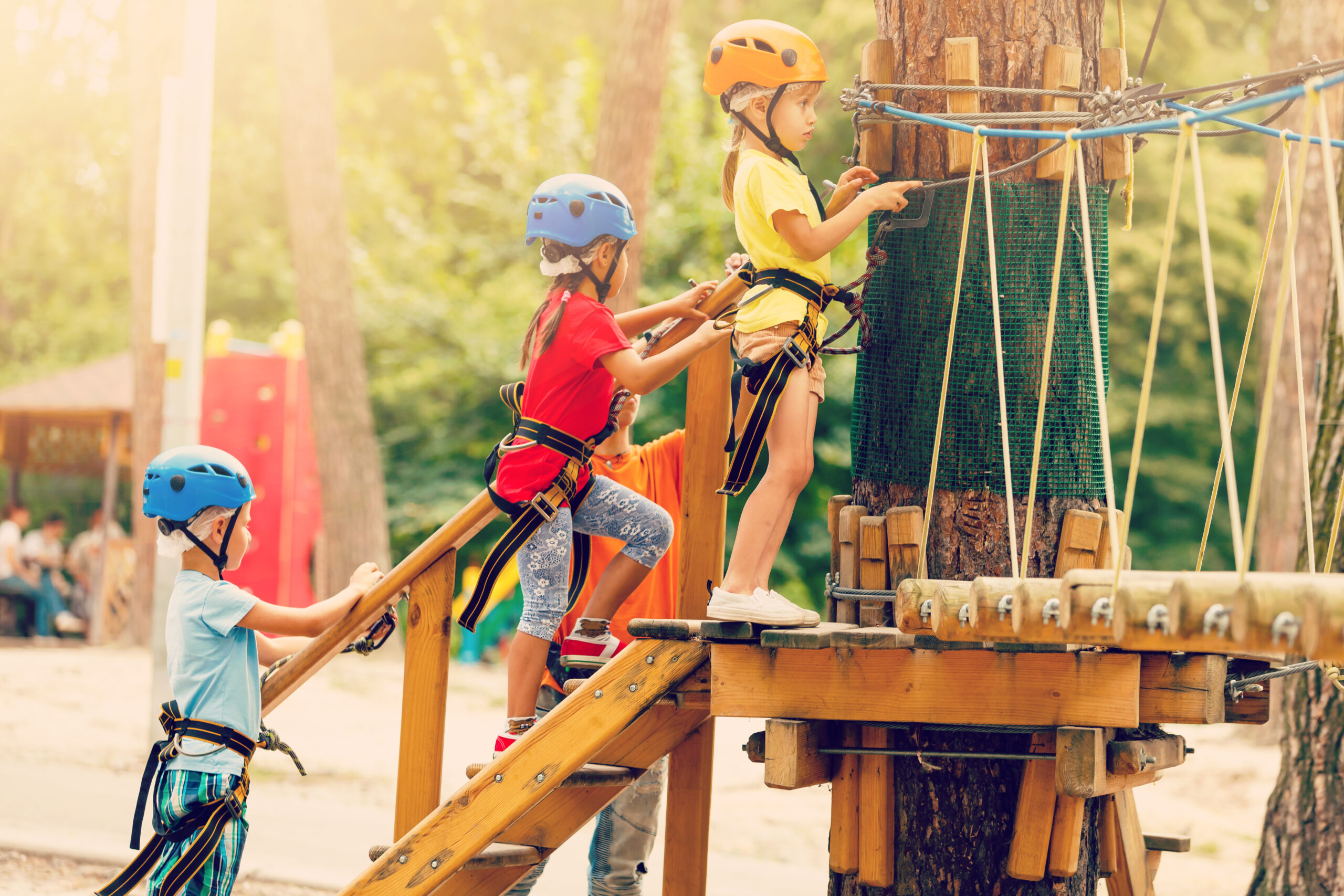 Three elementary-aged children stand in line on wooden steps waiting for their turn on a summer camp ropes course. Each child is wearing a safety helmet and harness connected to overhead rope lines while preparing for the outdoor activity.