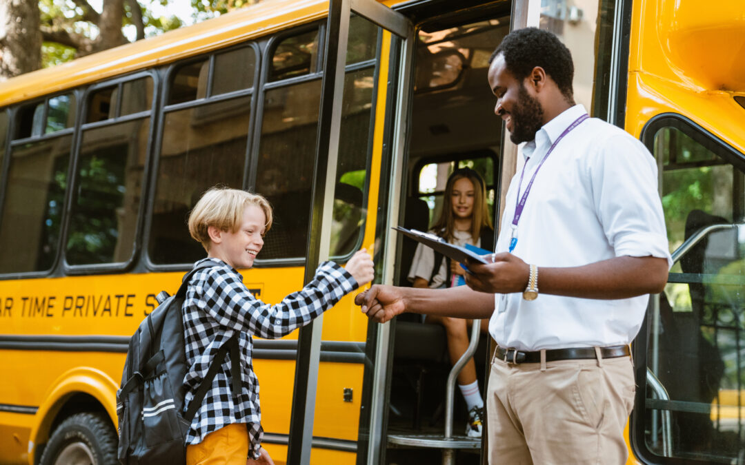 Teacher holding a clipboard welcomes a student with a fist bump before the student boards a yellow school bus with classmates already seated inside.