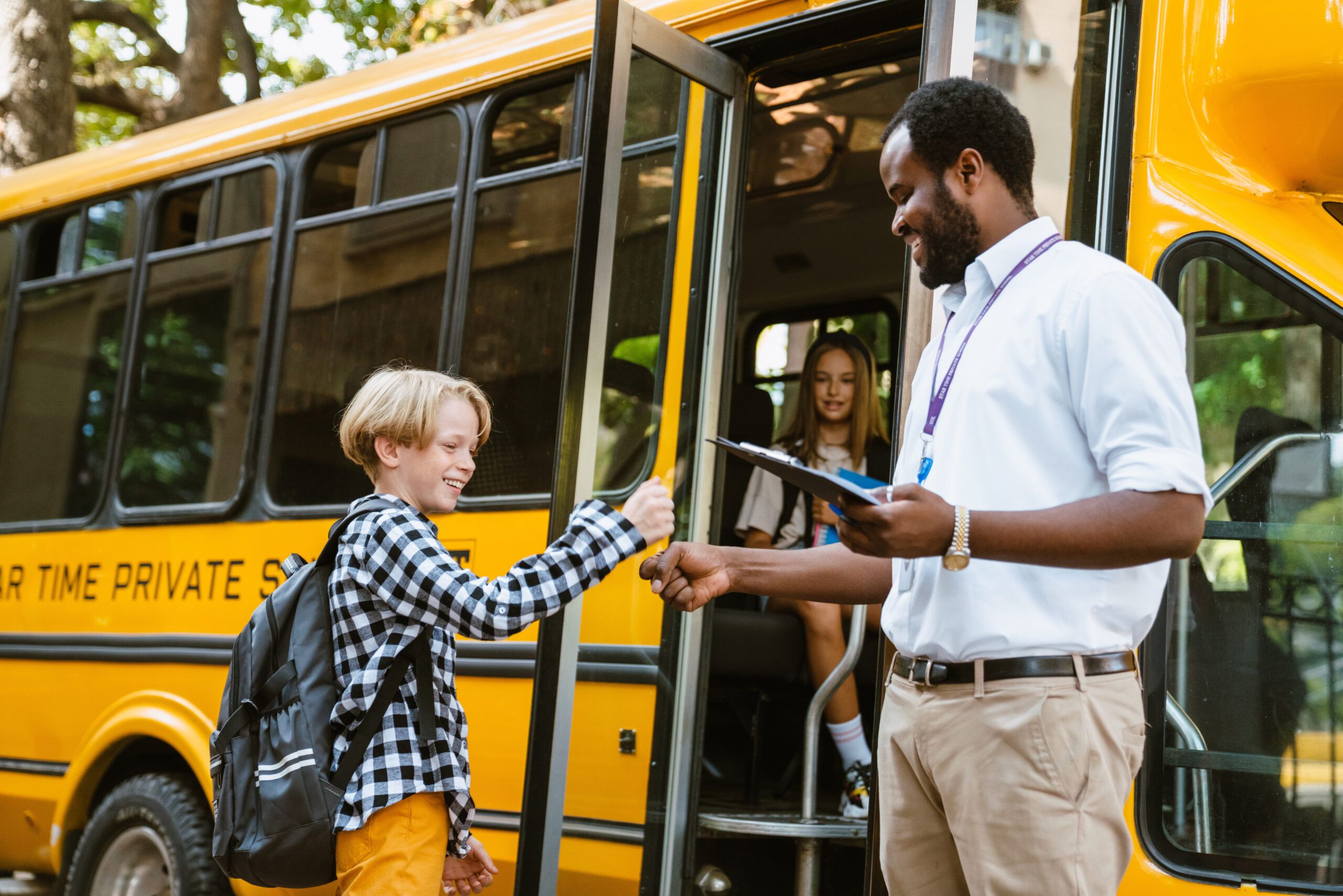 Teacher holding a clipboard welcomes a student with a fist bump before the student boards a yellow school bus with classmates already seated inside.