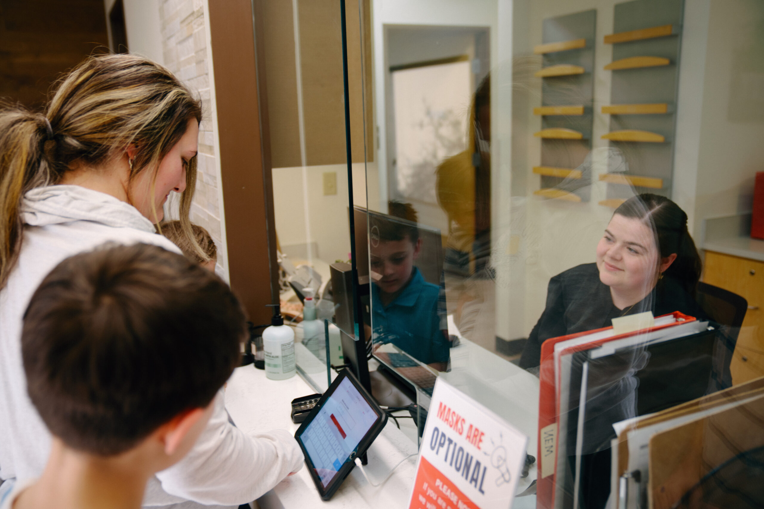 Mother and sons using a tablet to check in to an AFC Urgent Care clinic while the smiling receptionist greets them. Mother and sons using a tablet to check in to an AFC Urgent Care clinic while the smiling receptionist greets them.
